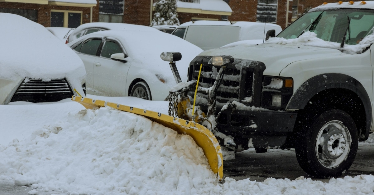 Commercial snow plow clearing snow in retail shopping center parking lot.